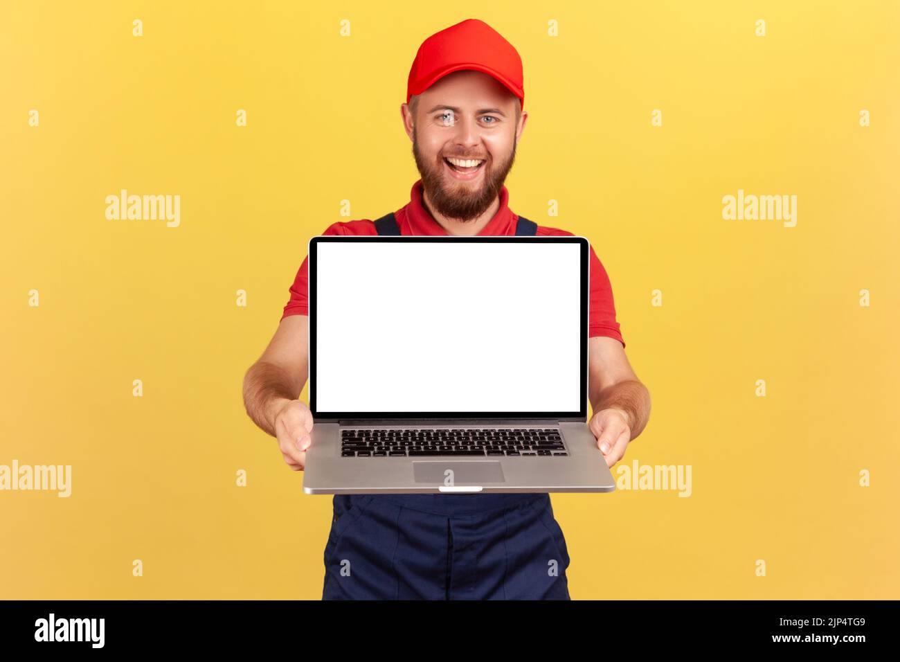 Portrait of delighted handsome worker man in red cap holding laptop, showing blank screen with copy space for advertisement and promotional text. Indoor studio shot isolated on yellow background. Stock Photo
