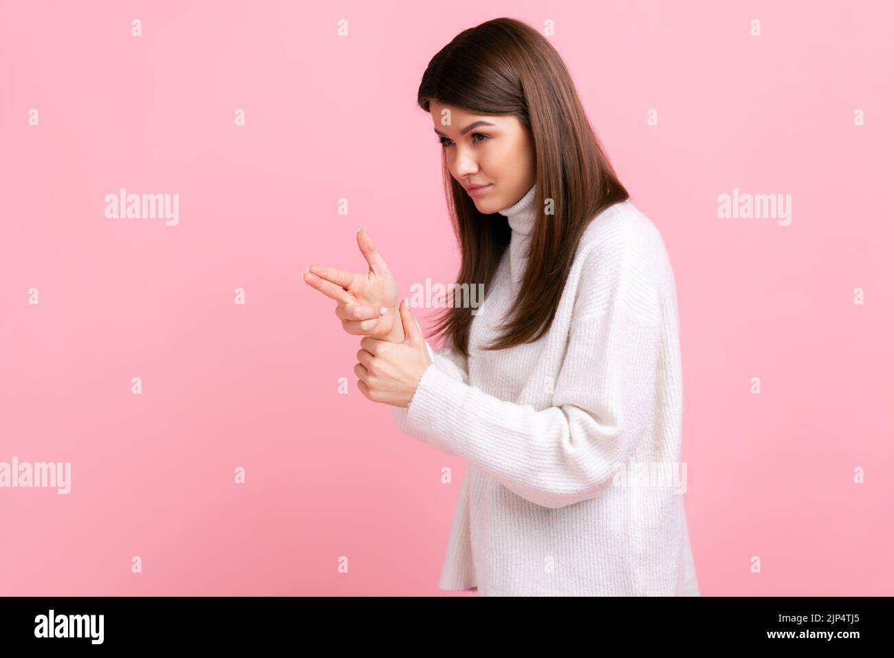 Dangerous criminal woman pointing finger gun, threatening to kill, shooting with weapon gesture, wearing white casual style sweater. Indoor studio shot isolated on pink background. Stock Photo