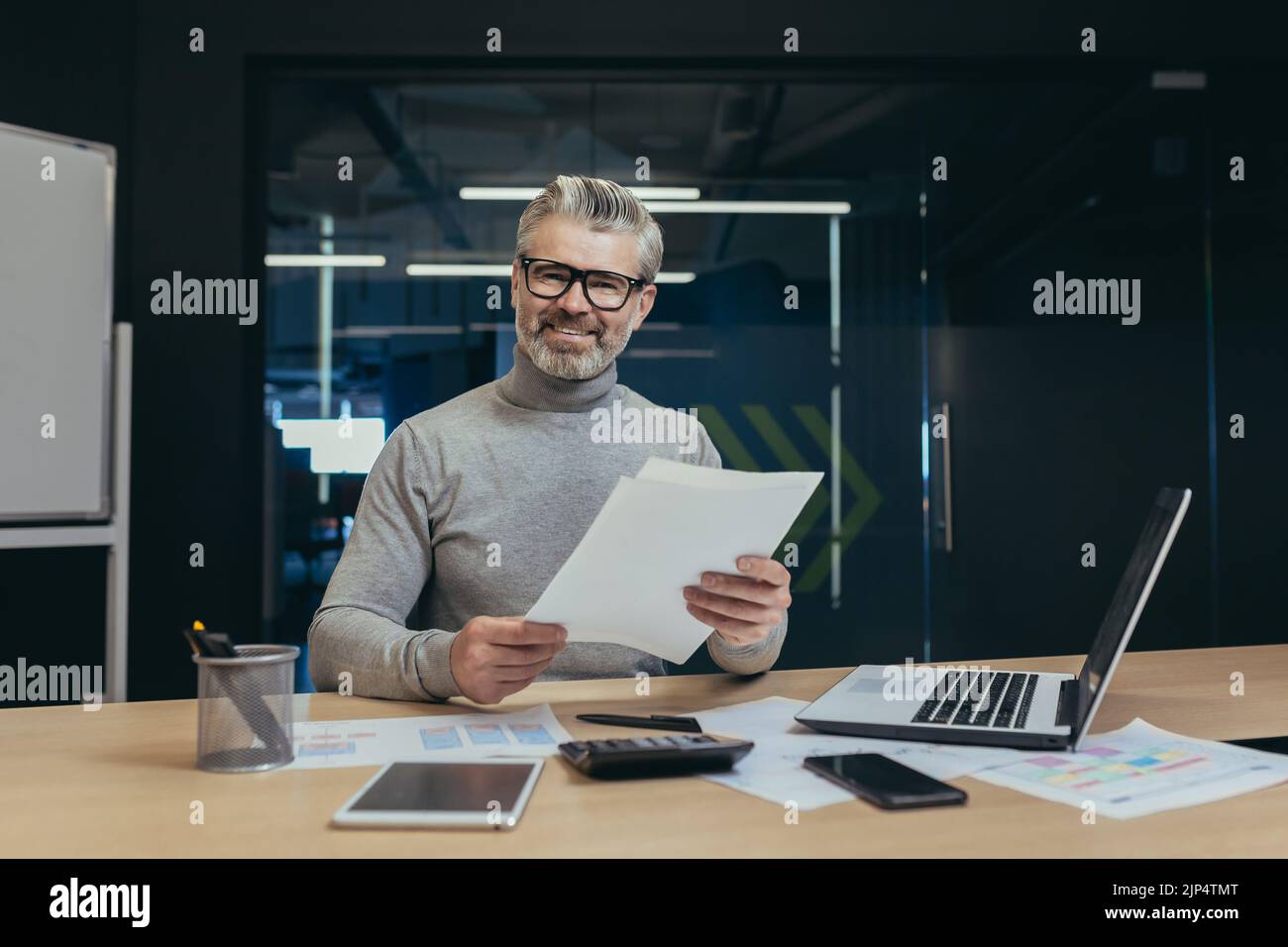 Portrait of mature experienced financier, man working with documents inside office building, businessman smiling and looking at camera holding documents and bills, boss working using laptop Stock Photo