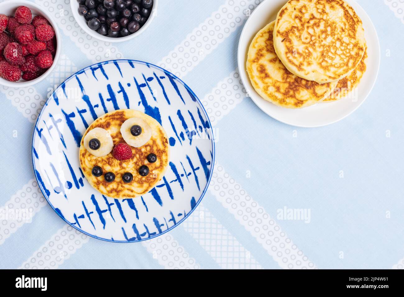 American pancakes decorated like smile and happy faces with raspberries, blueberries and banana. Food for kids, playful and creative. Top view. Stock Photo