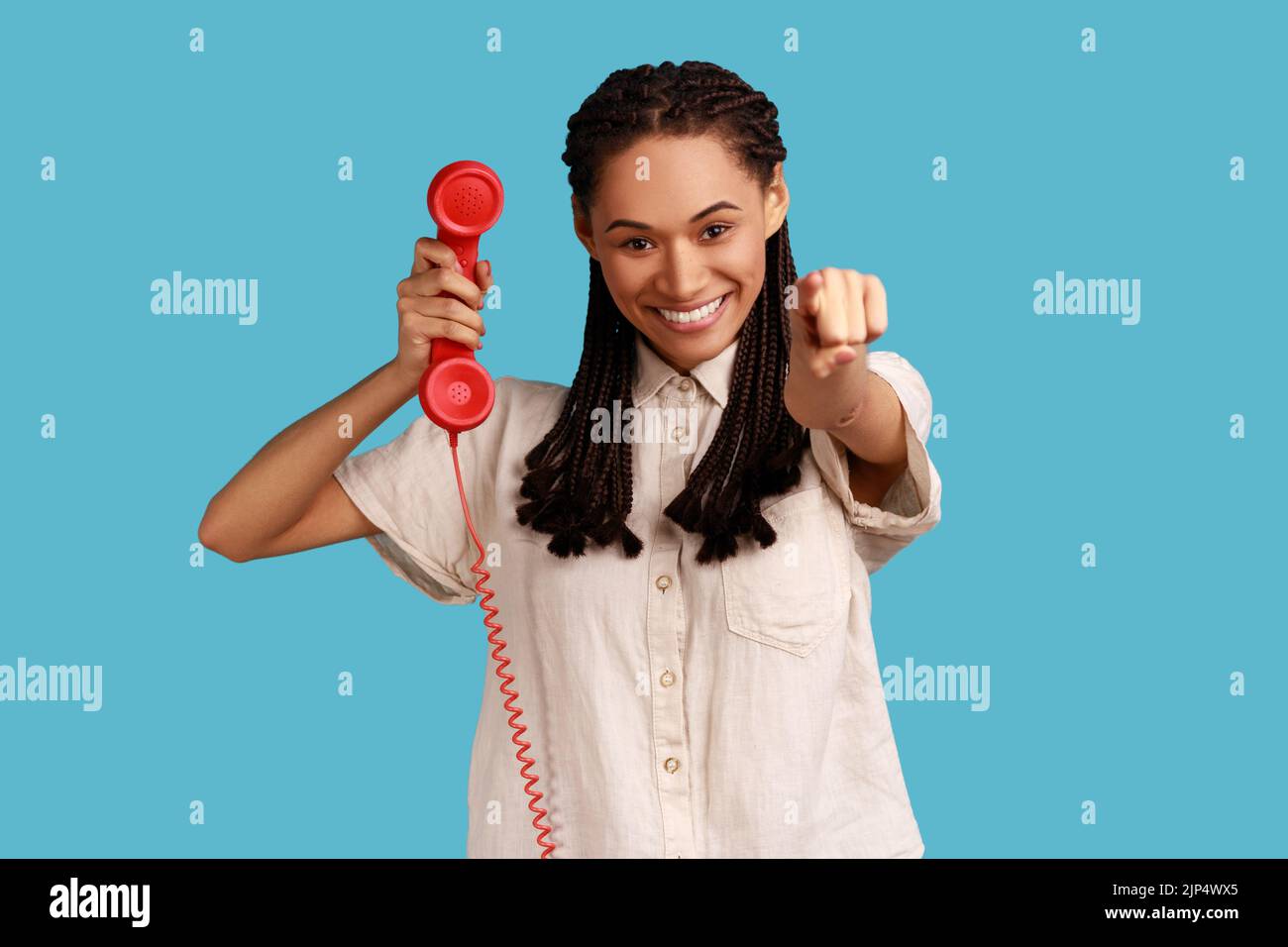 Beautiful woman with black dreadlocks pointing finger to camera, holding handset of red vintage landline phone, answering calls, wearing white shirt. Indoor studio shot isolated on blue background. Stock Photo
