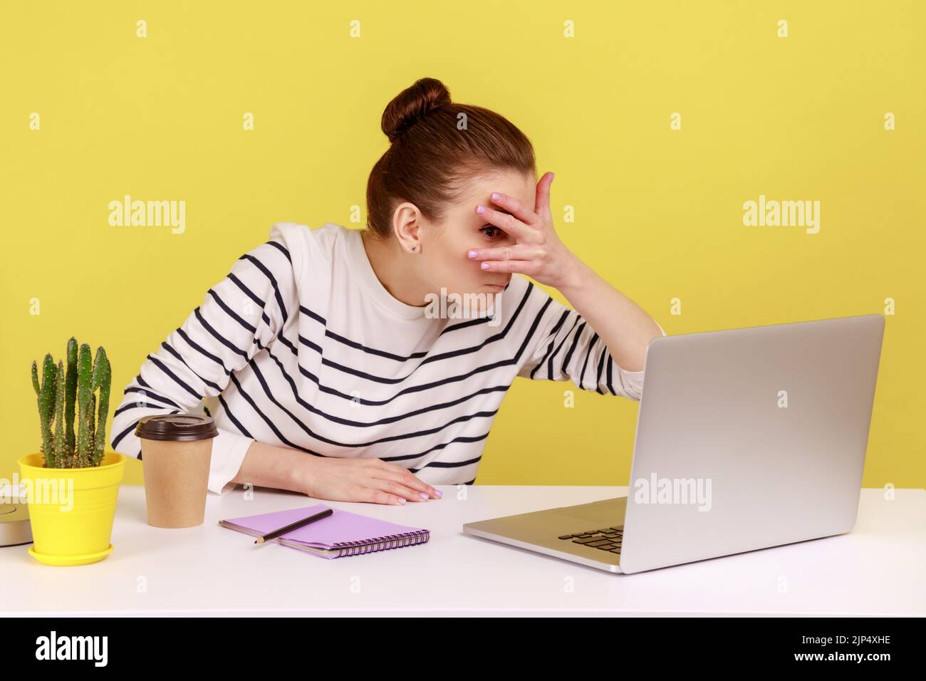 Curious woman manager sitting at workplace, spying, looking through fingers at laptop screen, peeking secret gossip in office. Indoor studio studio shot isolated on yellow background. Stock Photo