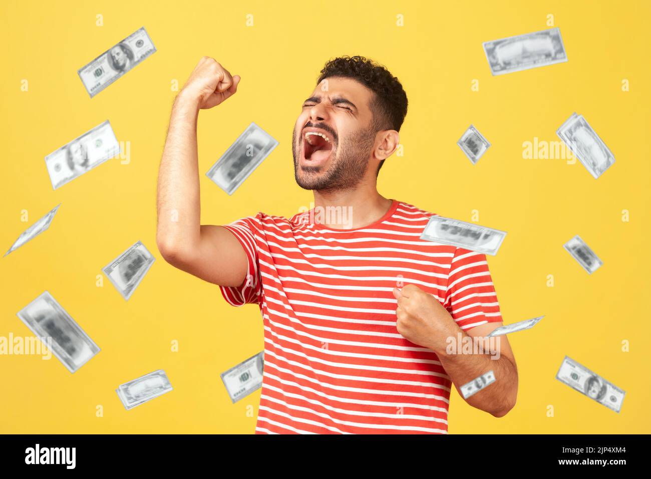 Money rain, winner and rich. Ecstatic happy celebrating motivated man standing with raised fists and shouting for joy, winner excited for success. dollars falling. indoor isolated on yellow background Stock Photo