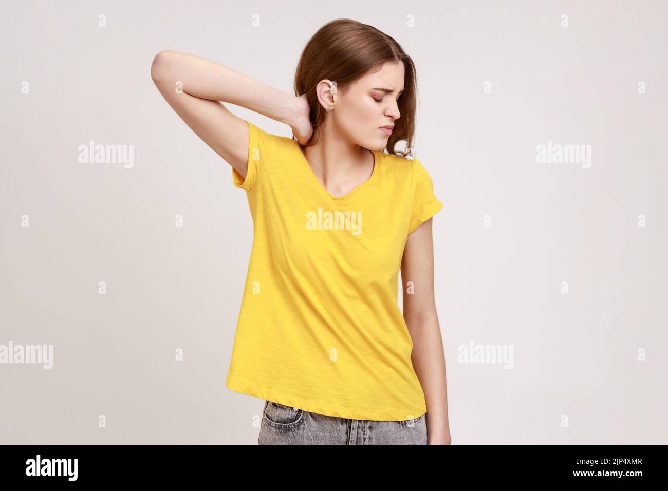 Shoulders and neck pain. Portrait of tired upset teen girl in yellow casual style T-shirt standing massaging neck to relieve pain, muscle strain in back. Indoor studio shot isolated on gray background Stock Photo