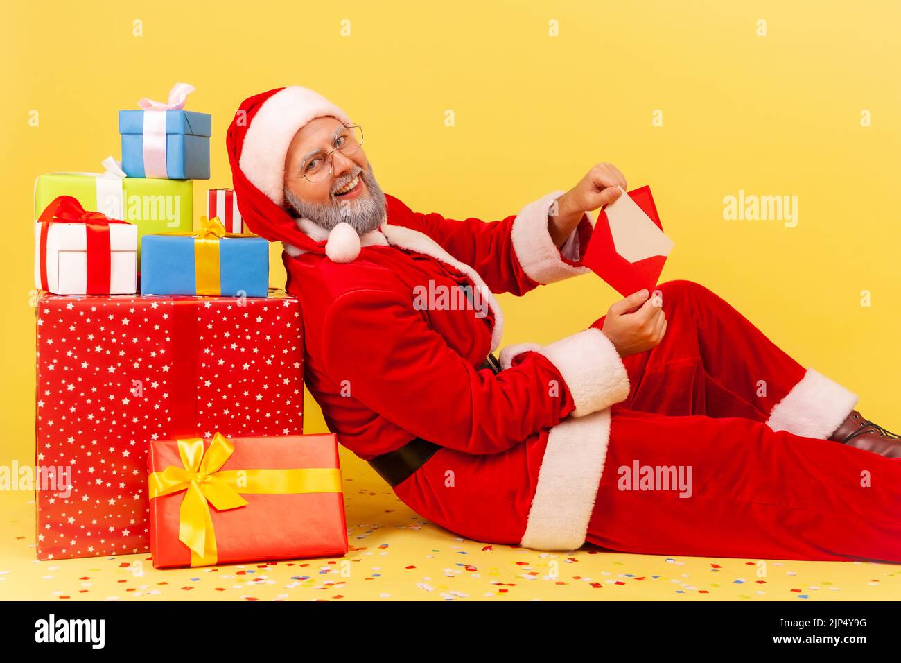 Profile portrait of elderly man with gray beard in santa claus costume sitting surrounded with Christmas boxes, holding envelope and smiling to camera. Indoor studio shot isolated on yellow background Stock Photo