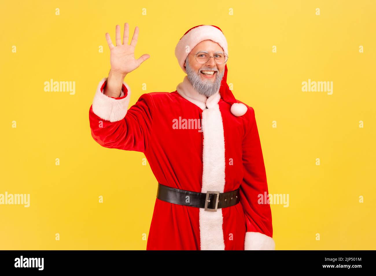Positive good looking elderly man with gray beard wearing santa claus costume waving hand, greeting somebody, looking at camera with smile. Indoor studio shot isolated on yellow background. Stock Photo