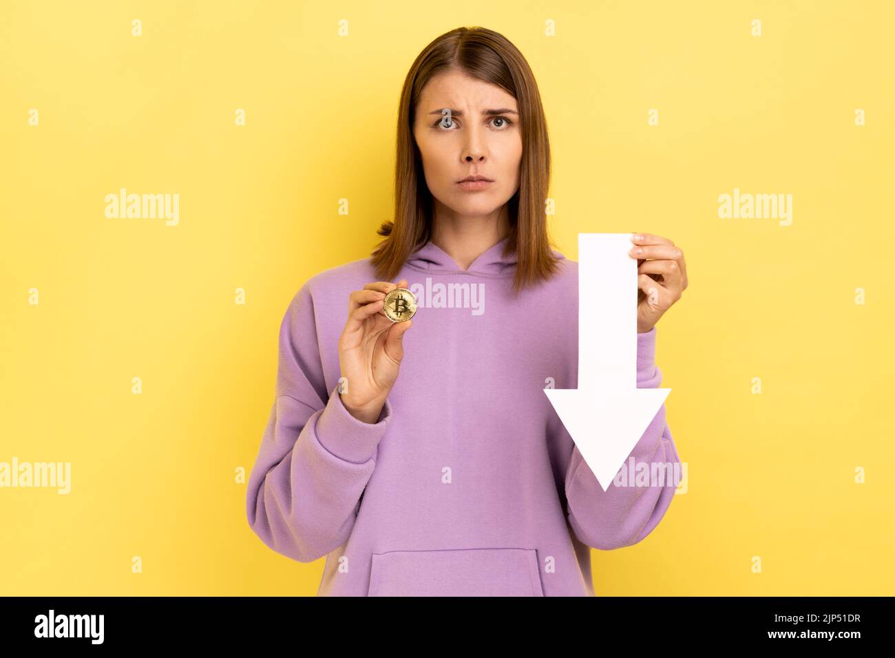Portrait of unhappy upset woman showing bitcoin and white arrow pointing down, expressing negative emotions, wearing purple hoodie. Indoor studio shot isolated on yellow background. Stock Photo