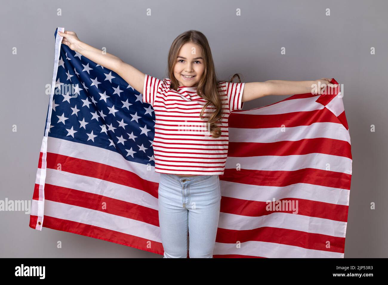 Portrait of winsome little girl wearing striped T-shirt holding huge american flag and yelling happily, celebrating national holiday. Indoor studio shot isolated on gray background. Stock Photo