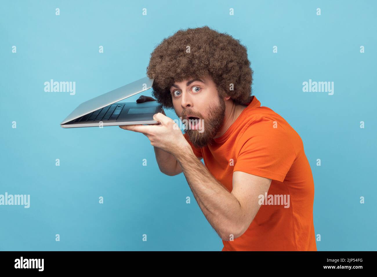Side view of surprised man with Afro hairstyle wearing orange T-shirt sees shocking information on somebody notebook, spying, looking at camera. Indoor studio shot isolated on blue background. Stock Photo