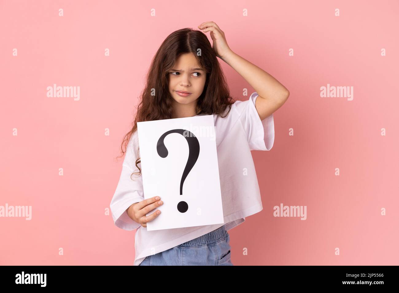 Portrait of little girl wearing white T-shirt holding paper with question mark over, thoughtful, face thinking about question, very confused idea. Indoor studio shot isolated on pink background. Stock Photo