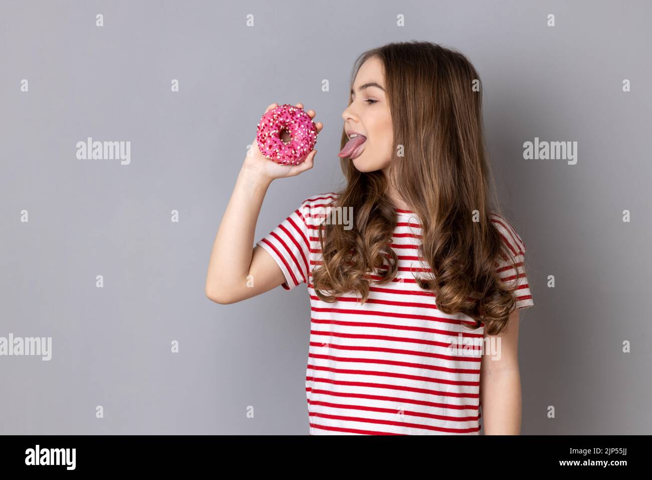 Portrait of hungry little girl wearing striped T-shirt standing licking doughnut and showing tongue out, sweet sugary confectionary. Indoor studio shot isolated on gray background. Stock Photo