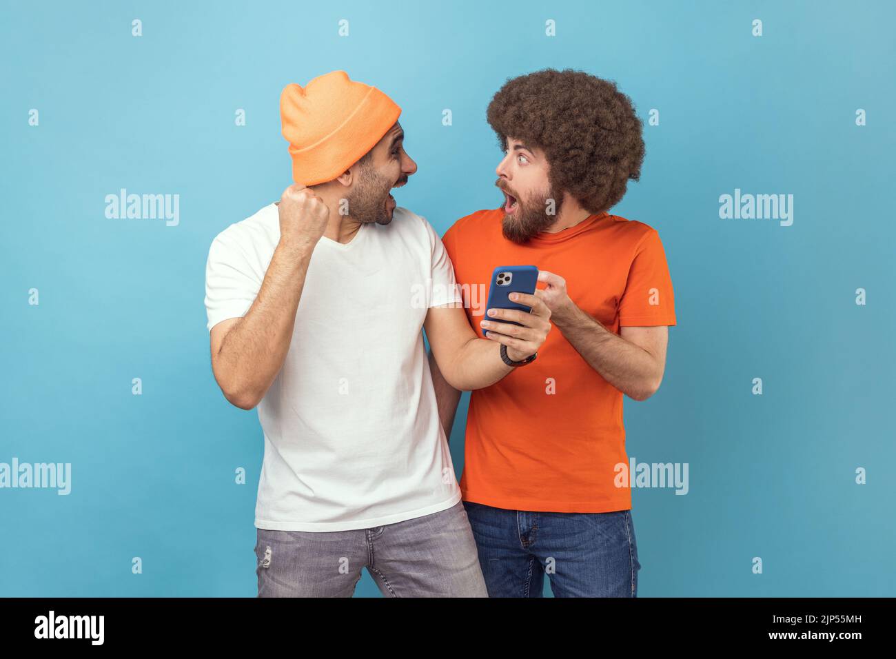 Portrait of two excited amazed young adult hipster men standing with cell phone and celebrating success, clenched fists, looking at each other. Indoor studio shot isolated on blue background. Stock Photo