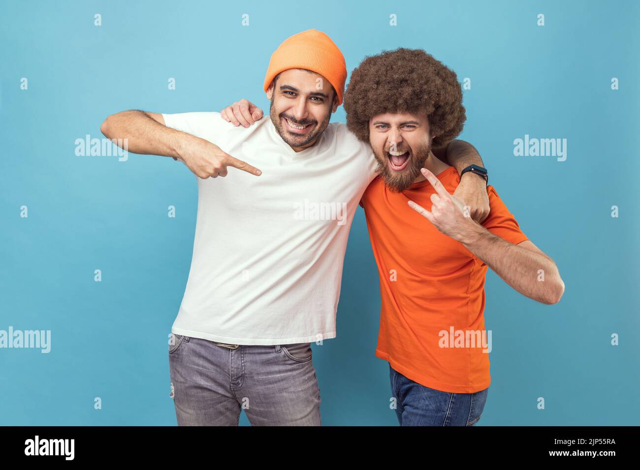 Portrait of two men hugging each other, guy with Afro hairstyle showing rock and roll sign hand gesture, male in beanie hat pointing at his friend. Indoor studio shot isolated on blue background. Stock Photo