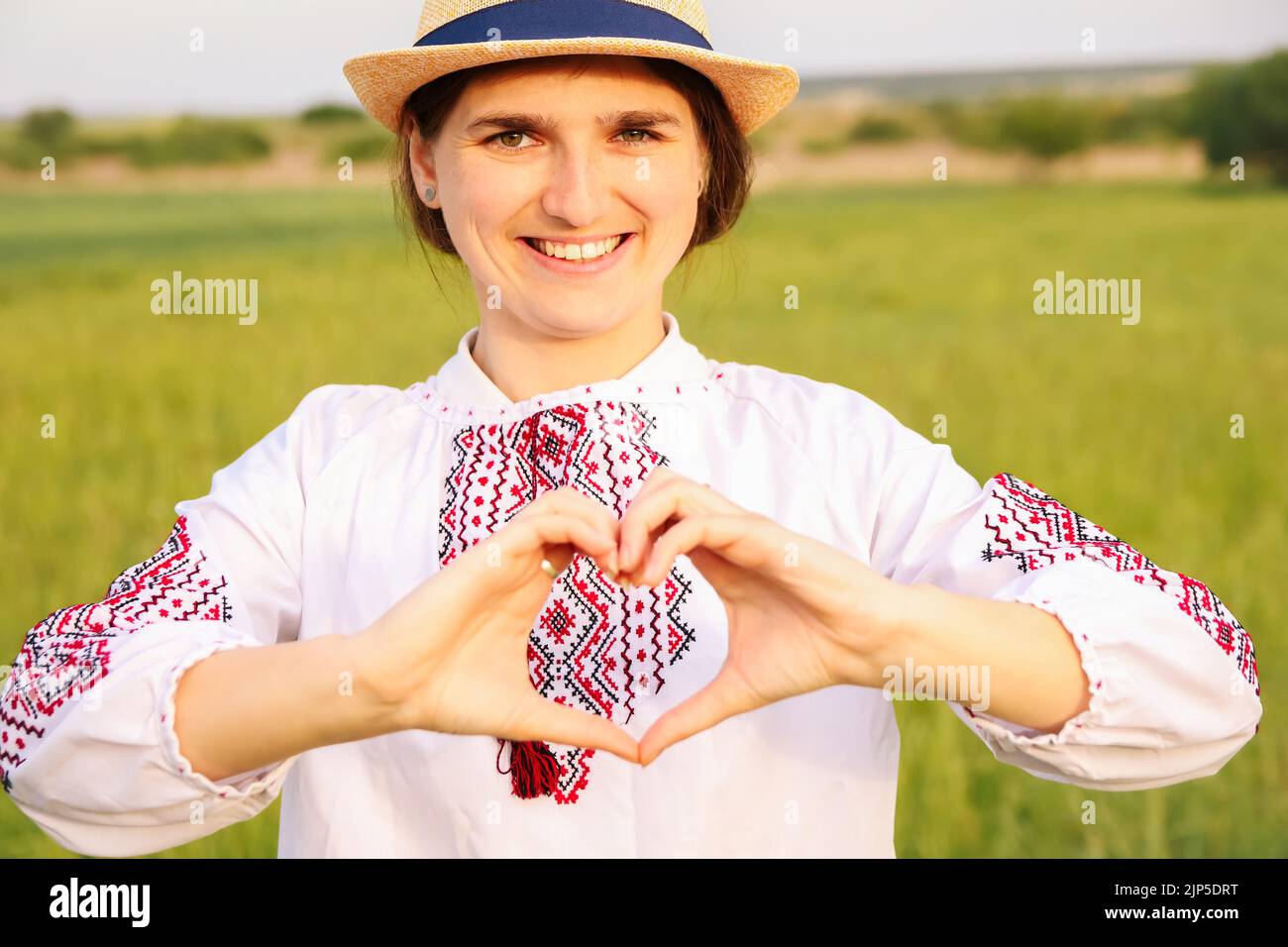 Defocus young ukrainian woman portrait. Meadow nature background. Showing sign love Ukraine. Smiling ukrainian girl showing gesture love. Vyshyvanka Stock Photo