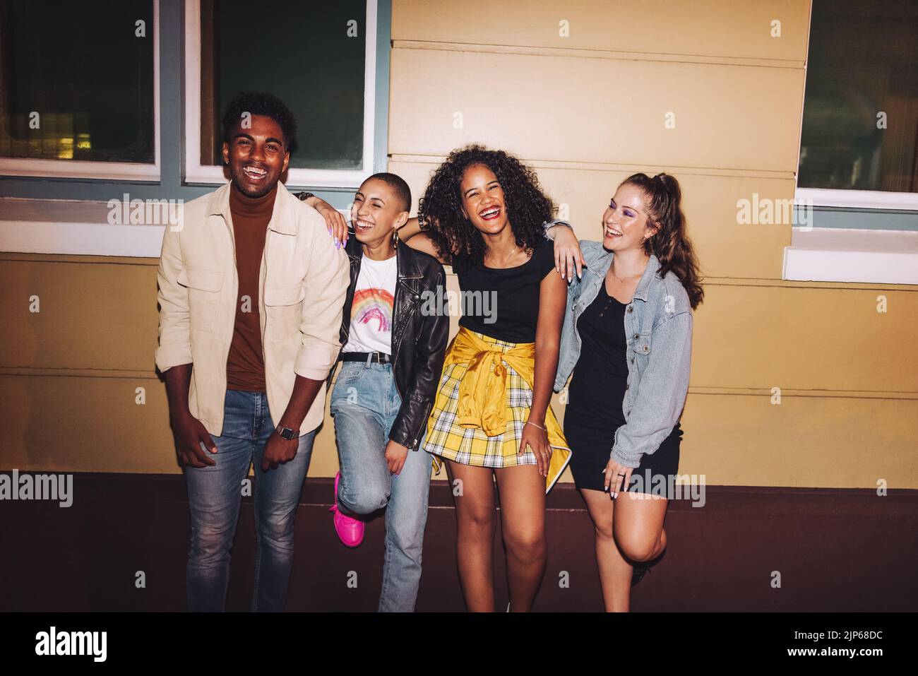 Cheerful friends leaning against a wall at night. Group of multicultural young people smiling happily while standing together outdoors. Carefree frien Stock Photo