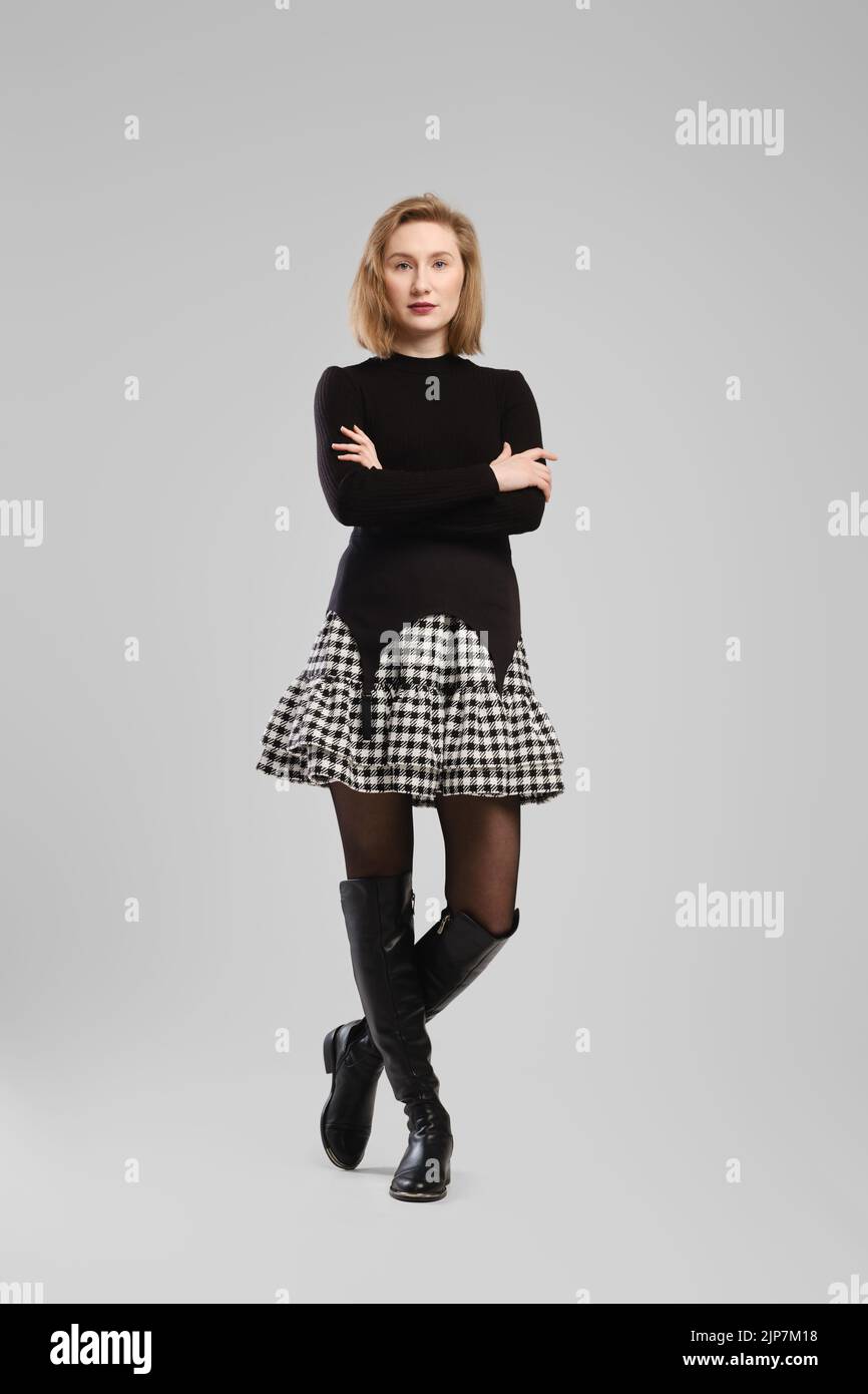 Full length portrait of young woman in turtleneck and suspender belt over little skirt posing in studio with crossed arms over grey background Stock Photo