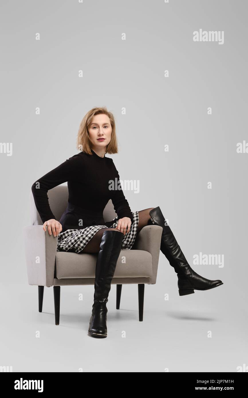 Trendy woman in turtleneck and suspender belt over little skirt sitting in armchair in studio over grey background Stock Photo