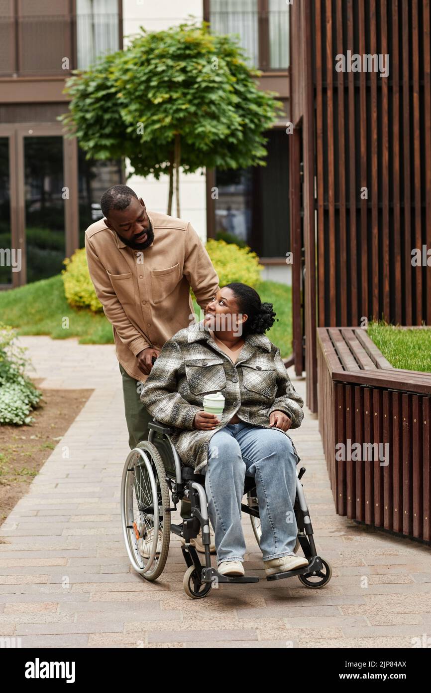 Vertical full length portrait of black couple with young woman in wheelchair enjoying walk in city garden and looking at each other Stock Photo