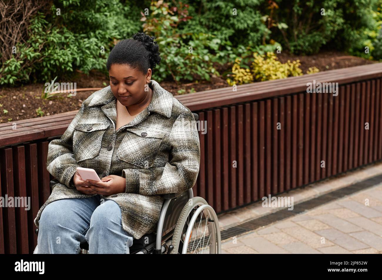 Portrait of black woman with disability using smartphone in city garden setting and wearing coat, copy space Stock Photo