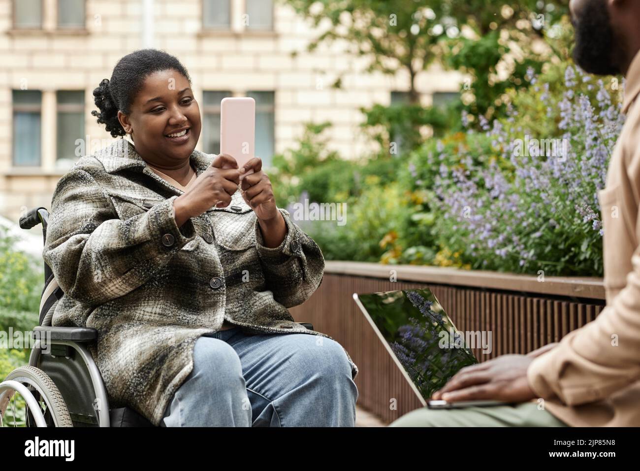 Portrait of smiling black woman in wheelchair taking mobile photo in city outdoors, copy space Stock Photo