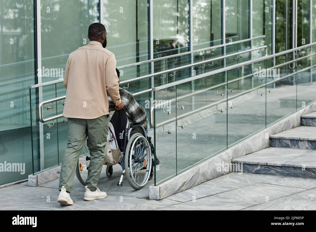 Back view of adult man assisting partner in wheelchair going up accessibility ramp in city, copy space Stock Photo