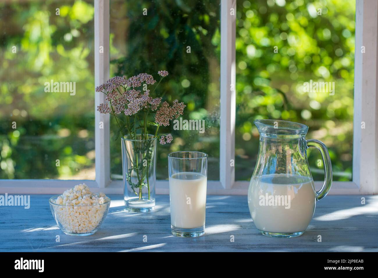 Milk in a jug in a glass and cottage cheese on the windowsill by the window of the house on a summer day near the garden, close up. Lots of dairy prod Stock Photo