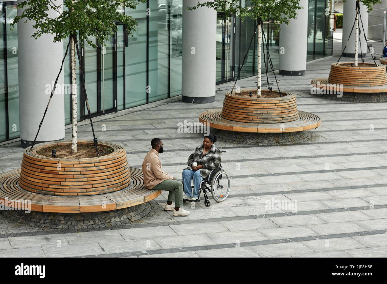 Wide angle view at adult black couple with partner in wheelchair chatting outdoors in city setting, graphic tiled floor, copy space Stock Photo