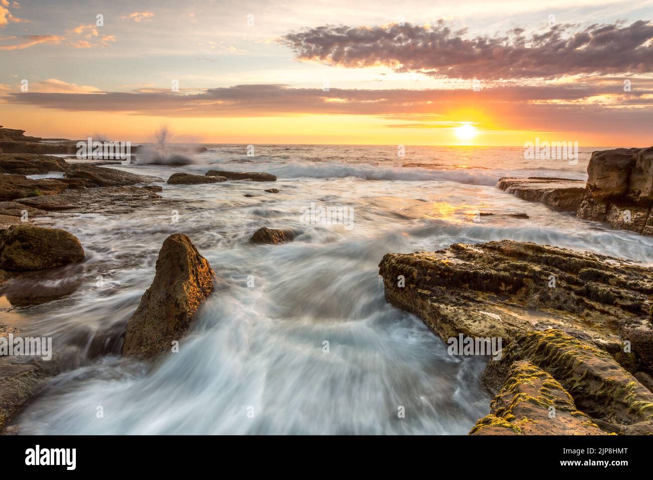 Beautiful coastal sunrise.  Waves flow over the rocky seashore and splash upwards in various directions,  A golden orange sunrise lights the pretty  s Stock Photo