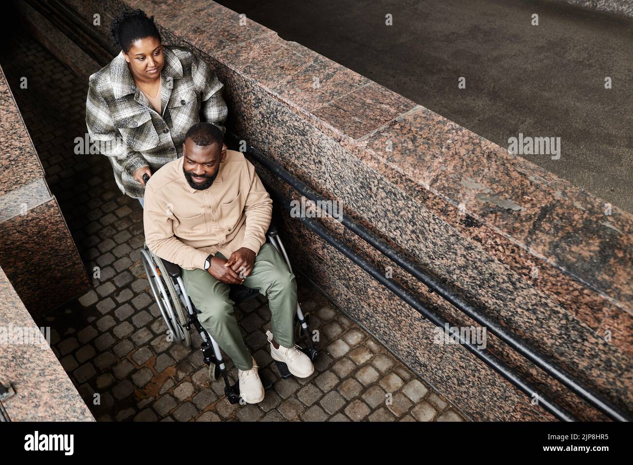 High angle portrait of black woman assisting partner in wheelchair going down ramp in city, copy space Stock Photo