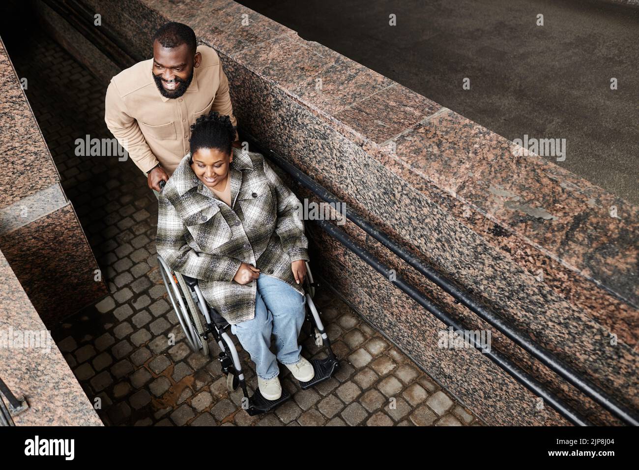 High angle portrait of black man assisting wife in wheelchair going down ramp in city, copy space Stock Photo