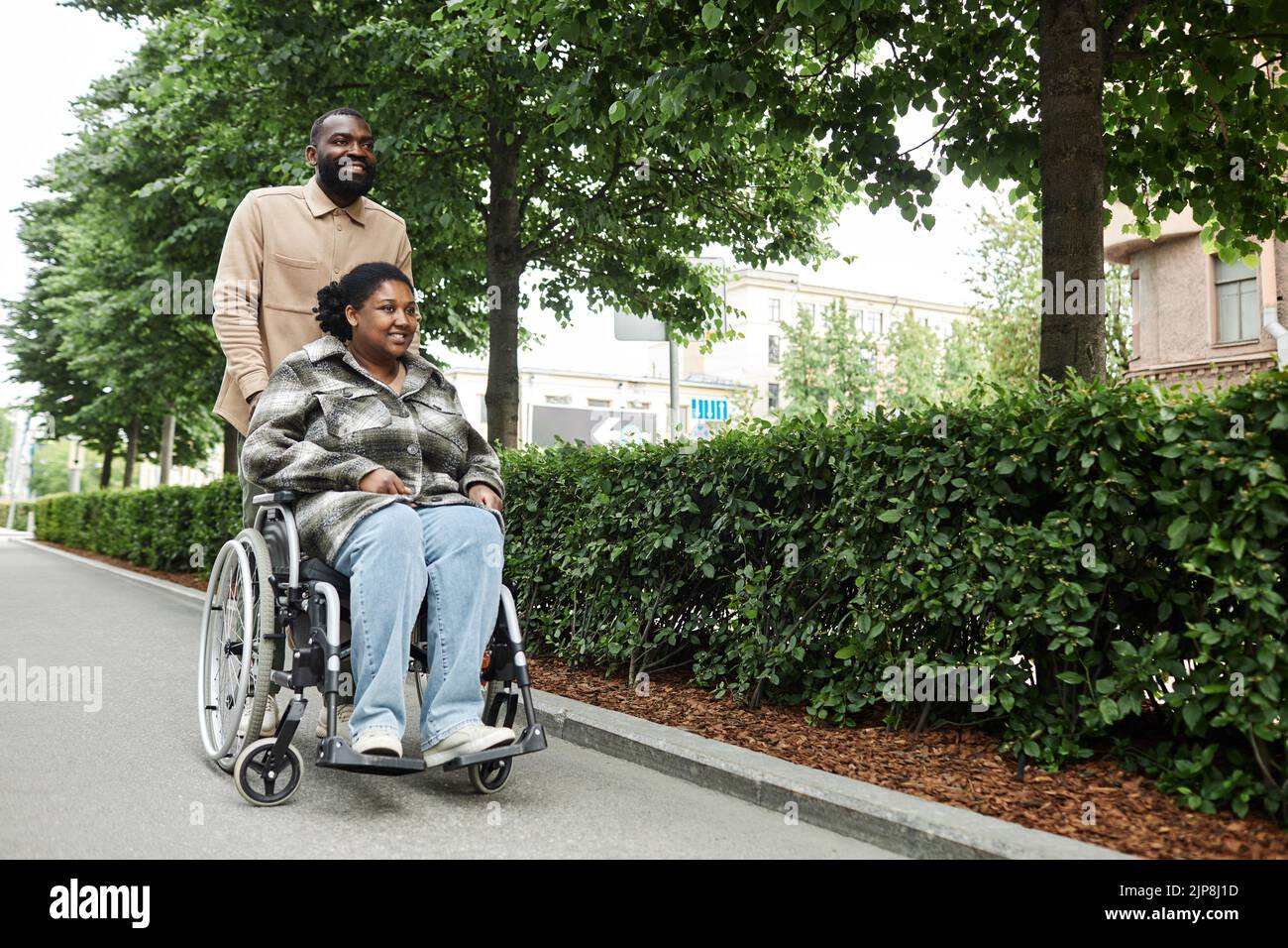 Full length portrait of happy adult couple with partner in wheelchair enjoying walk outdoors in city park, copy space Stock Photo