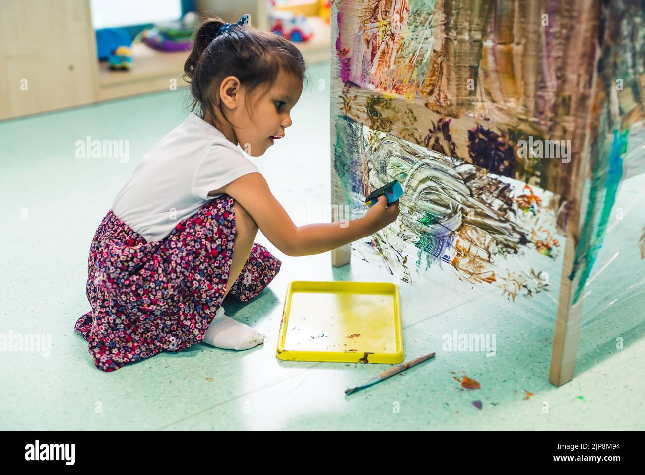 Cling film painting. Little girl toddler painting with a sponge and paints on a cling film wrapped all the way round the wooden shelf unit. Creative activity for kids development at the nursery school. High quality photo Stock Photo