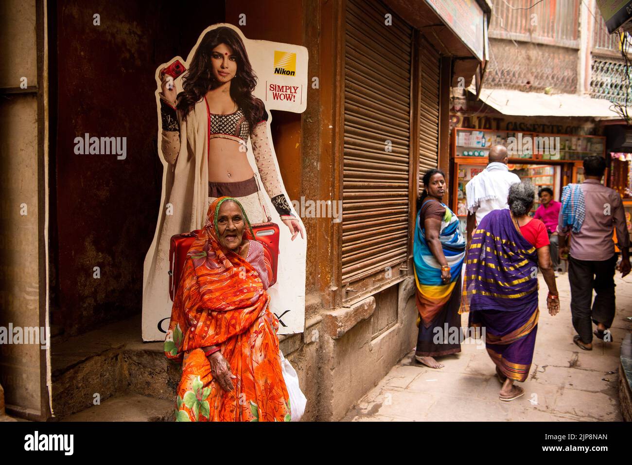 Old Indian woman wearing saree sitting below Nikon shop poster of bikini model in narrow lanes of Varanasi, Banaras, Benaras, Kashi, Uttar Pradesh, India Stock Photo