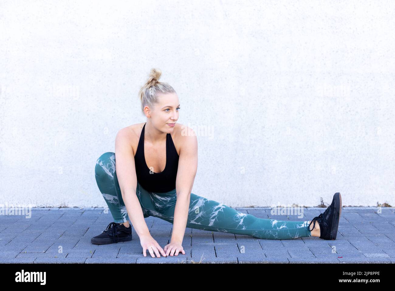 Beautiful young blonde fitness woman wearing black and blue sports clothes stretching and warming up quadriceps and glutes, buttocks, on legs in morning. Sporty woman doing warm up workout by a wall. High quality photo Stock Photo
