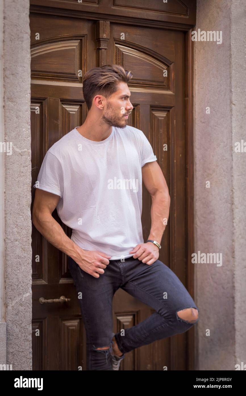 Self assured male model wearing white t shirt and black ripped jeans leaning on wooden door in street and looking away Stock Photo