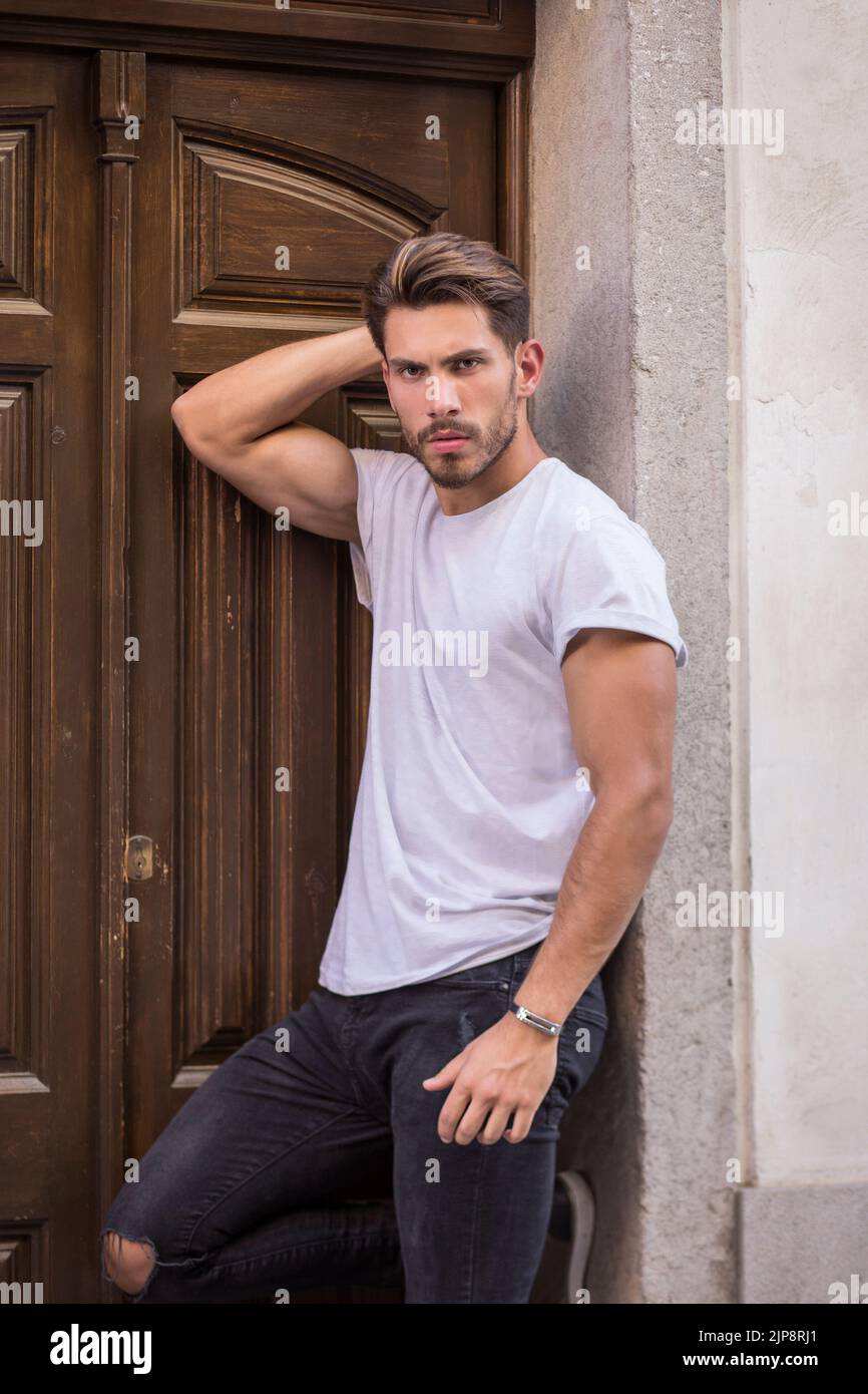 Self assured young male model in white t shirt and ripped jeans touching hair and looking at camera while standing near entrance door of building on s Stock Photo