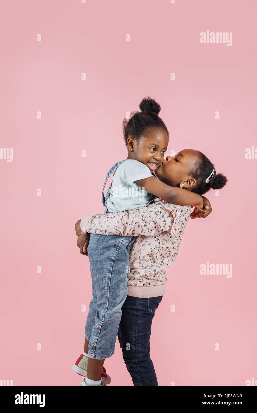 Two cute African girls sisters in casual summer outfits, hugging each other and one girl kissing other in her cheek on pink studio background, copy space. Stock Photo