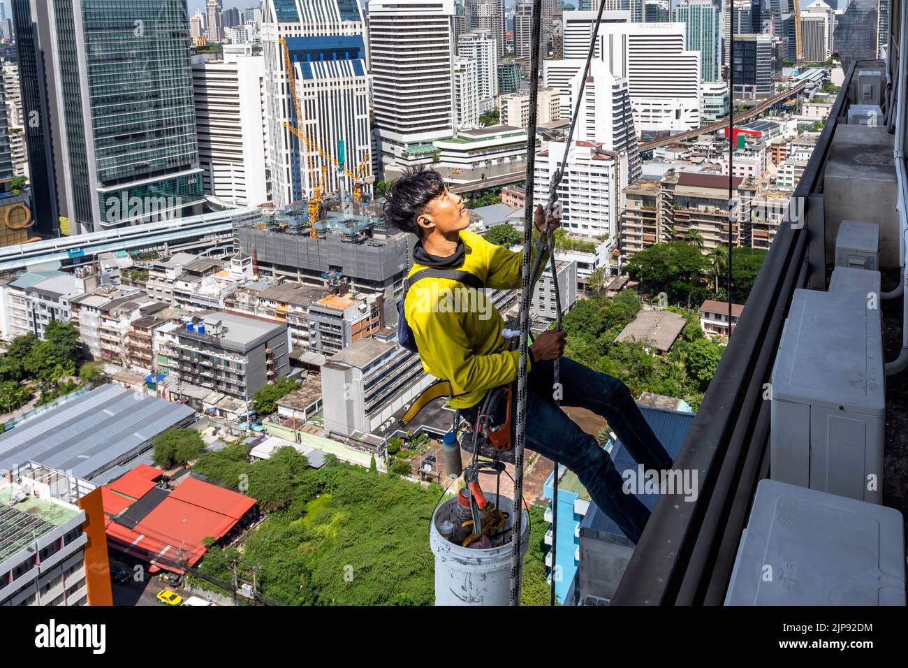 Thai worker using Bosun Chair to repair skyscraper, abseiling above Bangkok, Thailand Stock Photo