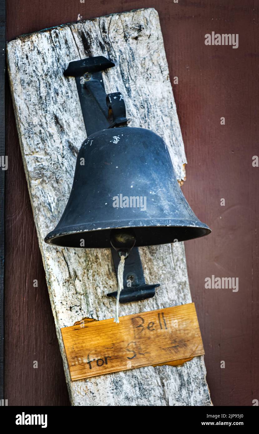 An old black iron bell attached to a rustic wooden plank with a string for ringing and a worn out sign underneath in any season in Pennsylvania Stock Photo