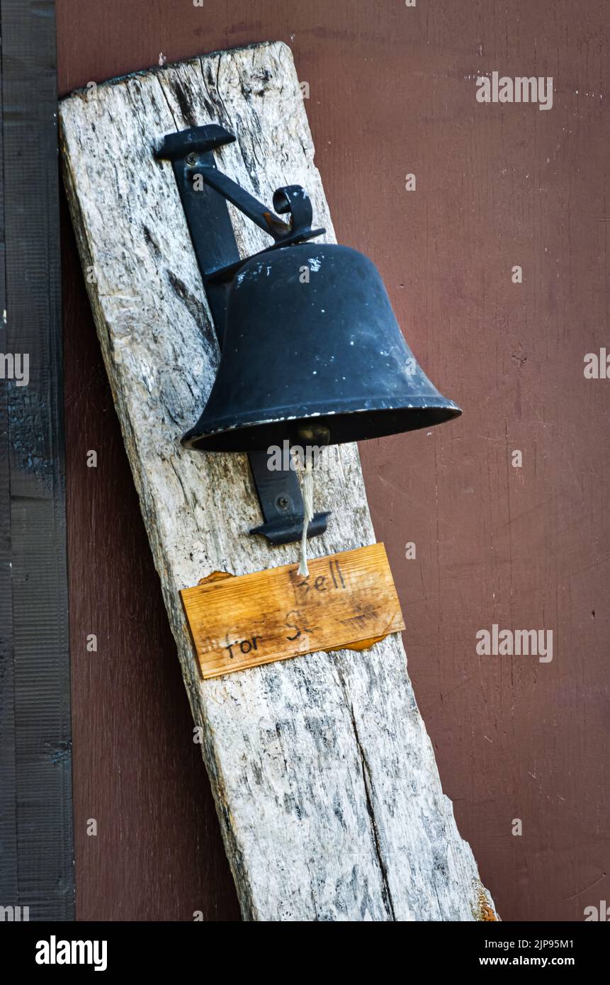 An old black iron bell attached to a rustic wooden plank with a string for ringing and a worn out sign underneath in any season in Pennsylvania Stock Photo