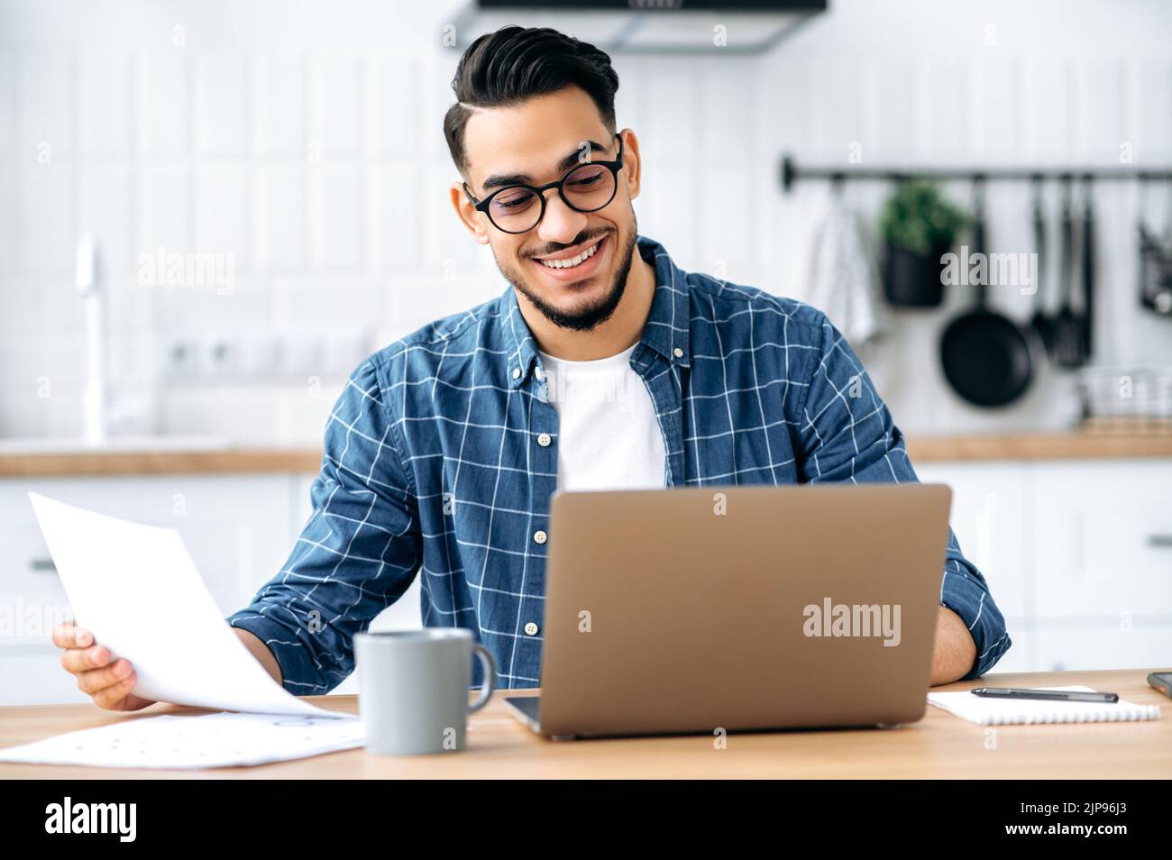 Work from home. Smart arabian or indian stylish man, freelancer, programmer, manager, working remotely, sit at a desk at home in the kitchen, working on a project uses laptop, studying documents,smile Stock Photo