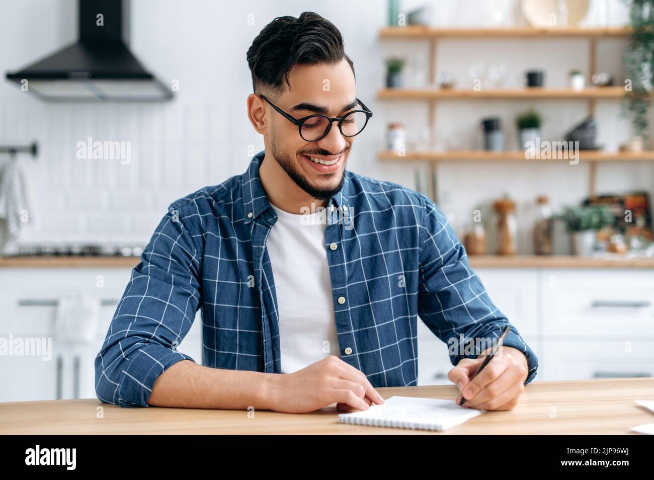 Clever excited Arabian or Indian modern guy, clever student or freelancer, with glasses, listening to an online webinar, lecture, writing down information, gain knowledge, smiling friendly Stock Photo