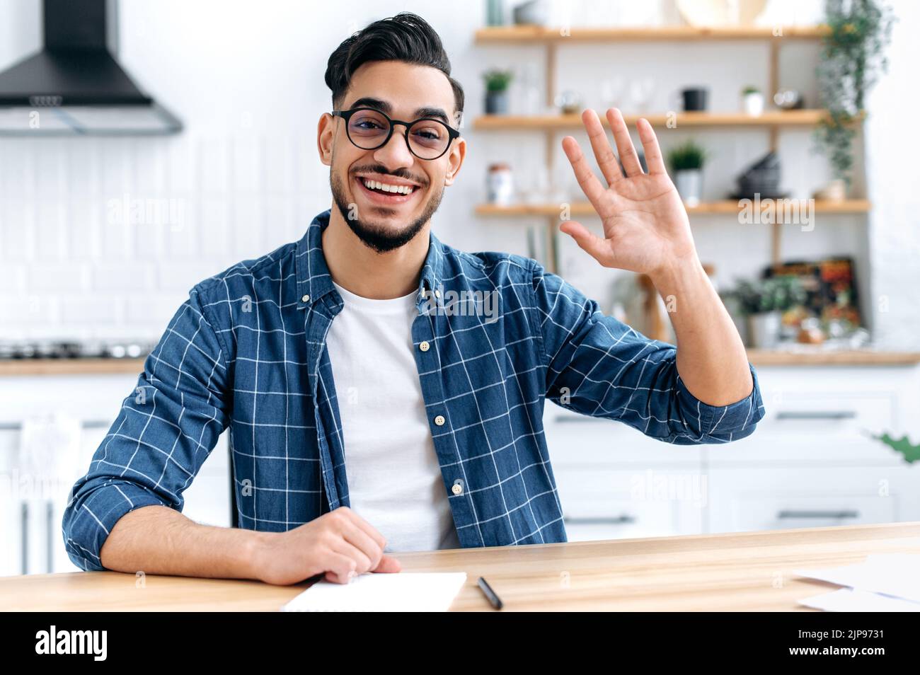 Confident positive indian or arabian man, sit at desk at home, talk on webcam, having video call or conversation with client or colleague, mentor record training course, waves hand, smiling friendly Stock Photo