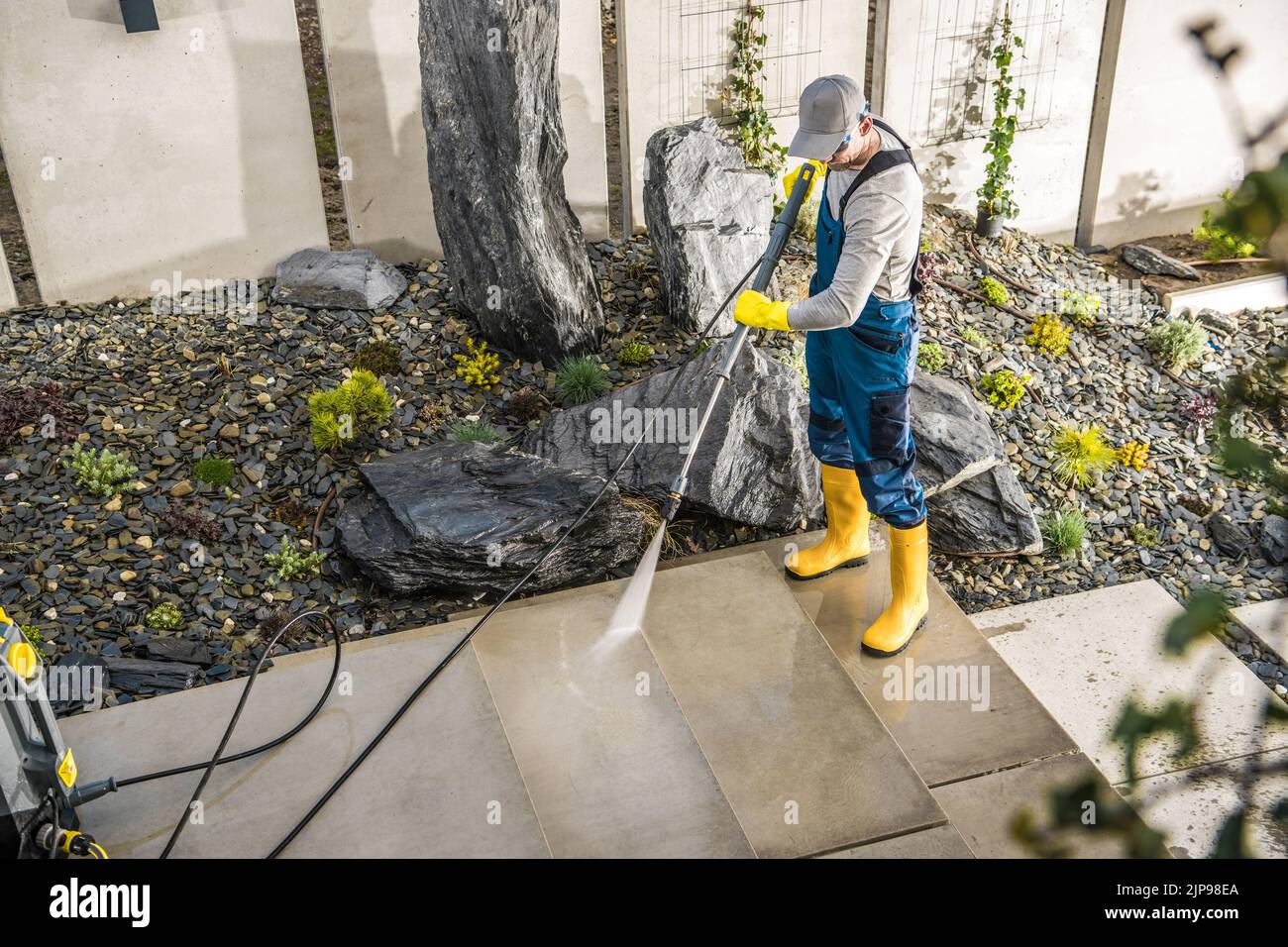 Caucasian Man in Protective Work Wear Cleaning Garden Paths with Pressure Washer. Decorative Stone and Gravel Aggregates in the Background. Spring Sea Stock Photo