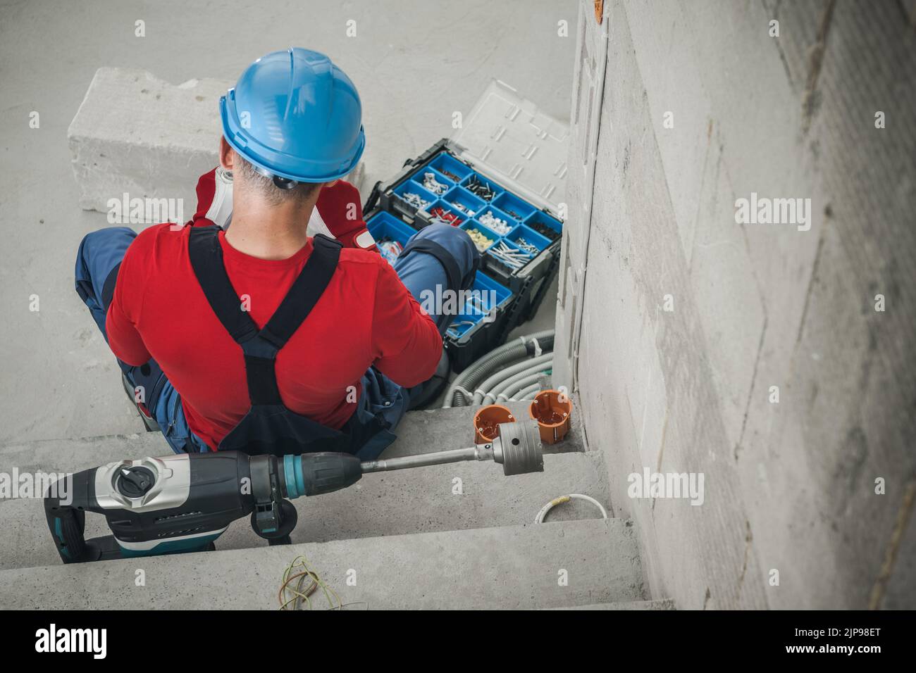 Caucasian Construction Worker Sitting in Front of His Opened Toolbox Preparing to Start Work. Power Drill with Hole Saw Bit Lying Alongside Him. Rear Stock Photo