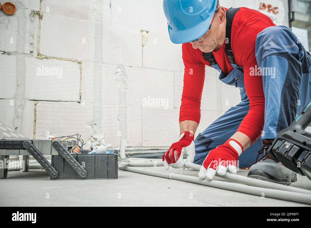 Caucasian Worker in Blue Hard Hat Installing Electric Cable System with Plastic Pipeline Coverage in Newly Built Residential Building. Industrial Them Stock Photo