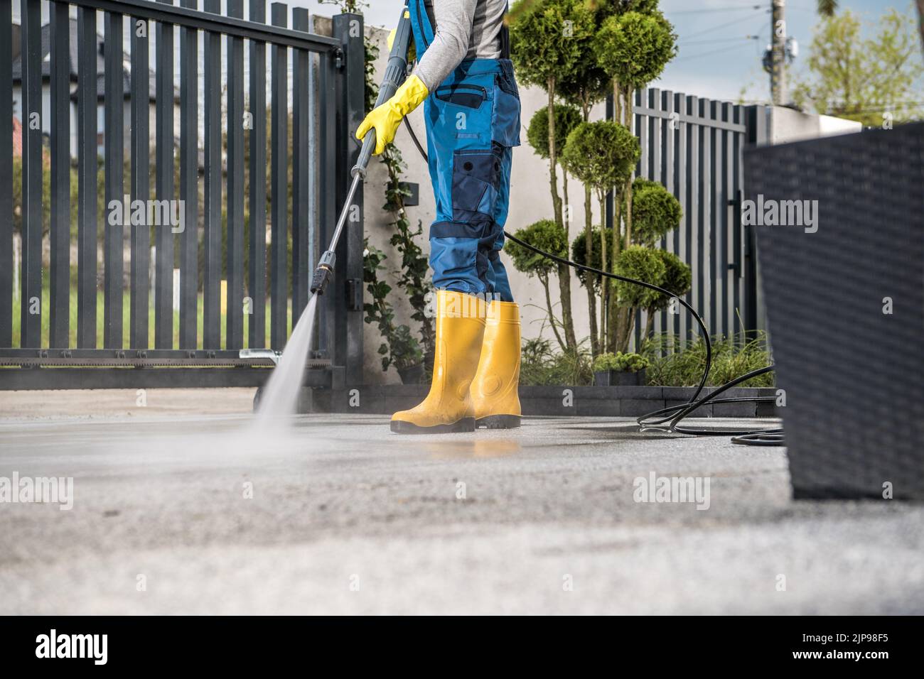 Man in Yellow Rain Boots and Work Uniform Pressure Washing Concrete Tiles of the Driveway to His House Behind the Closed Yard Entrance Gate. Home Surr Stock Photo