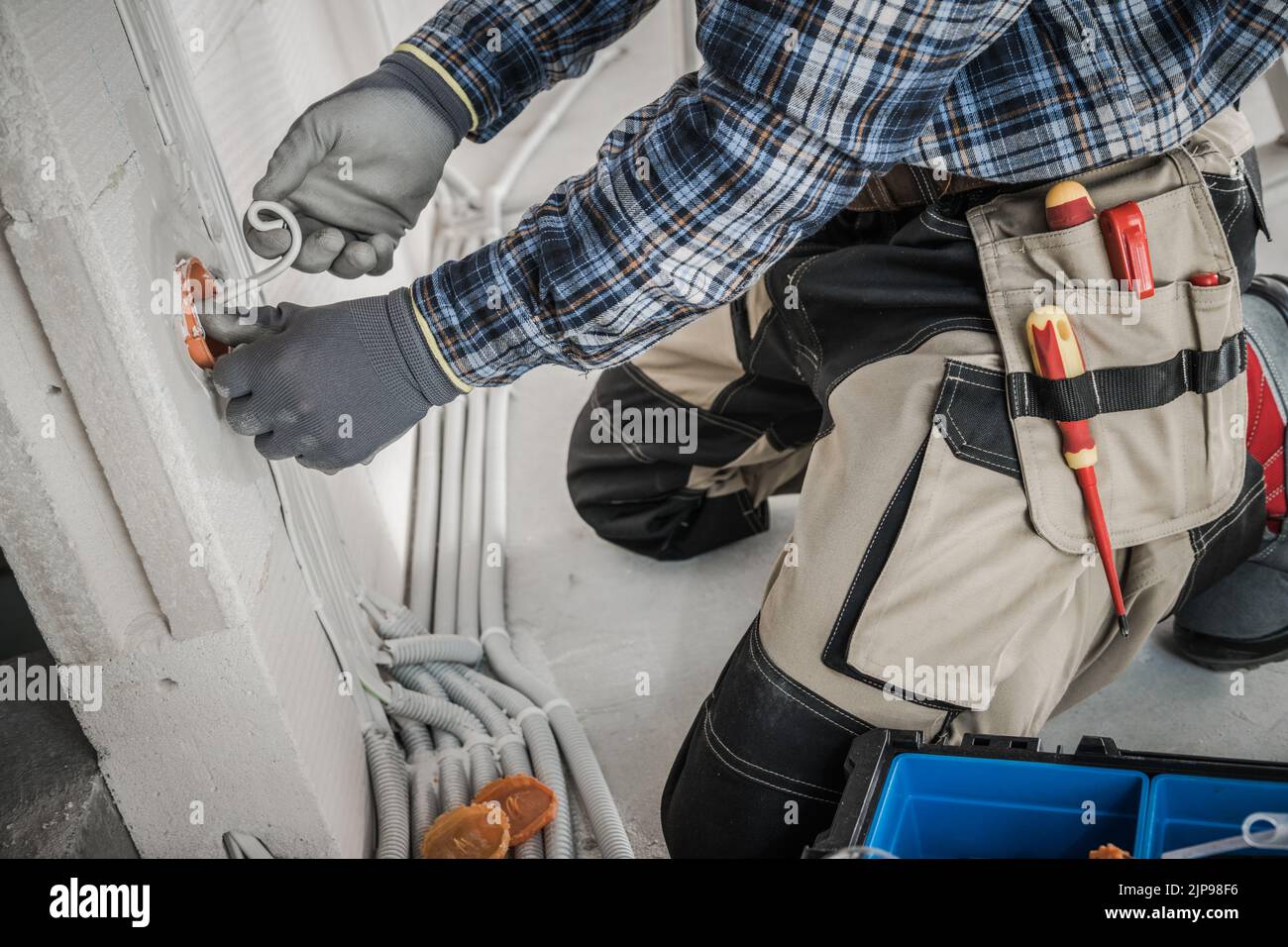 Professional Electrician in Protective Gloves and Tool Belt Working on Installing Power Sockets in the Wall. Plastic Pipeline in the Background. Indus Stock Photo
