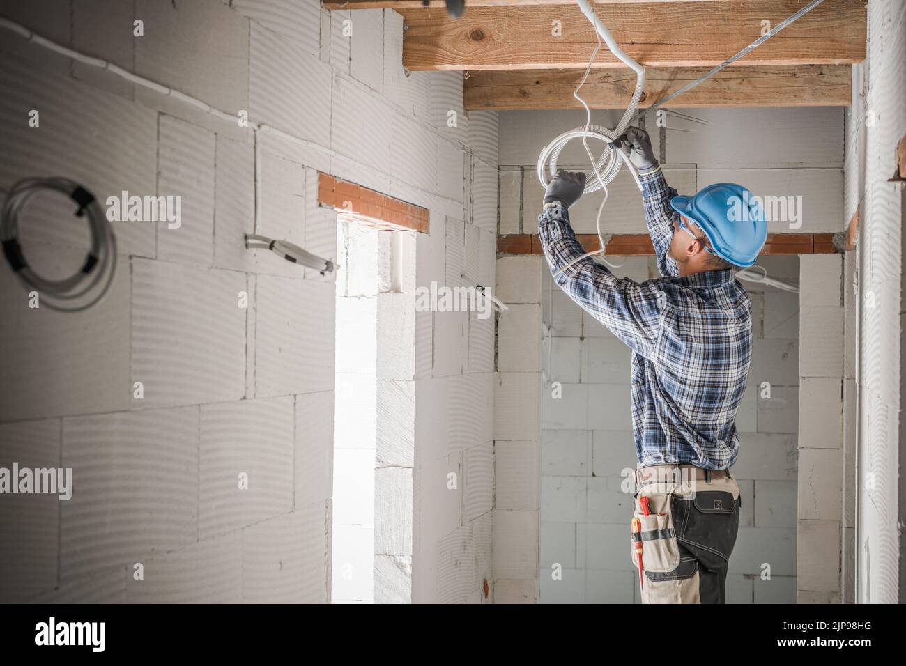 Caucasian Electrician Installing Power Line in Newly Built Residential Unit Pulling Cables Under the Ceiling. Construction Site in the Background. Ind Stock Photo