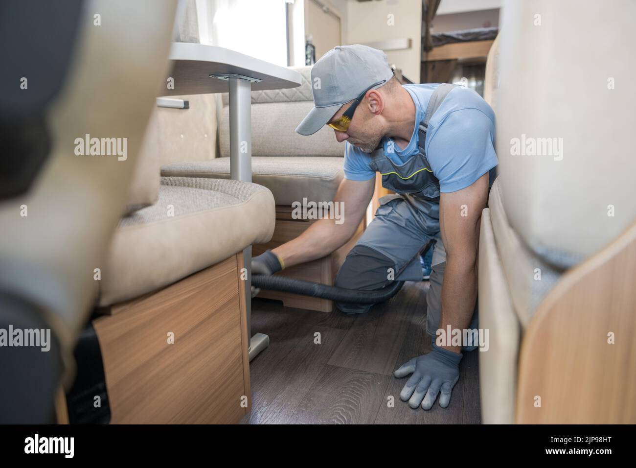 Recreational Vehicle Year-Round Care and Maintenance Services. Caucasian Worker Carefully Vacuuming Floor Under the Beige Sofa in Modern RV. Interior Stock Photo